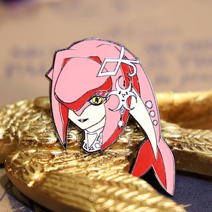 May include: A pink and red enamel pin featuring a stylized character with a large head, large eyes, and a long, flowing tail. The character is wearing a necklace with a pendant.