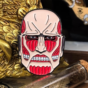 May include: A pin featuring a stylized Colossal Titan head from the anime series Attack on Titan. The pin has a light skin tone with red muscle details, a stern expression, and a black outline. The pin is set against a gold background.