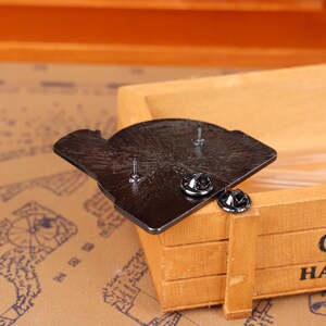 May include: A black enamel pin with two pin backs. The pin is in the shape of a stylized design. The pin is resting on a wooden box.
