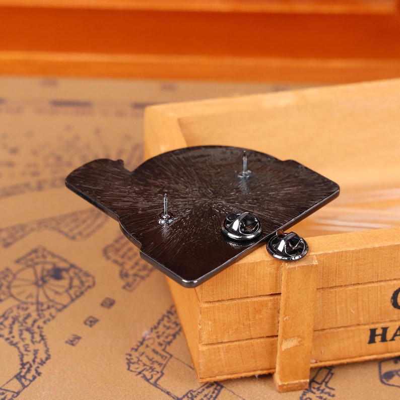 May include: A black enamel pin with two pin backs. The pin is in the shape of a stylized design. The pin is resting on a wooden box.