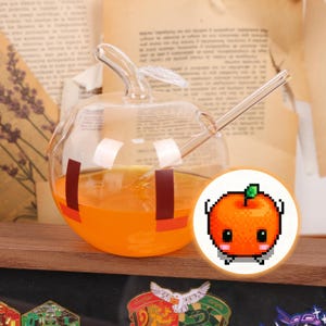 Könnte beinhalten: Ein klares, apfelförmiges Trinkglas mit orangefarbener Flüssigkeit, einem Glasstrohhalm und einer kleinen orangefarbenen Pixelkunstgrafik. Das Glas hat ein Stiel- und Blattdesign.