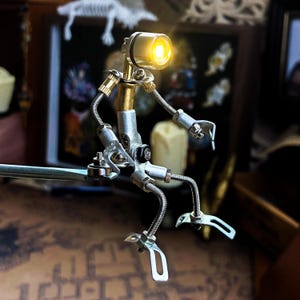 May include: A small, articulated robot made of metal and brass with a light on its head. The robot is sitting on a metal bar and has a silver and gold finish.