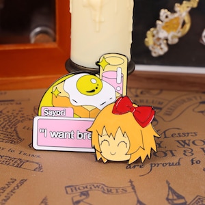 May include: Enamel pins featuring a cartoon character with a red bow, a fried egg on toast, and a drink. The pins have the text "Sayori" and "I want bre". The pins are colorful with yellow, pink, and orange accents.