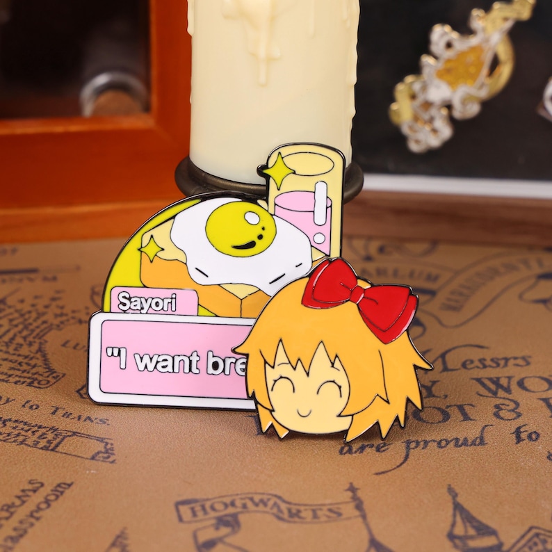 May include: Enamel pins featuring a cartoon character with a red bow, a fried egg on toast, and a drink. The pins have the text "Sayori" and "I want bre". The pins are colorful with yellow, pink, and orange accents.