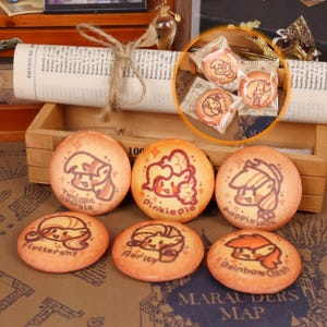 May include: A collection of round, orange-colored cookies, each decorated with a cartoon character design and a name. The cookies are arranged on a wooden tray, with some individually wrapped in clear plastic. The names include "Twilight Sparkle" and "Pinkie Pie".