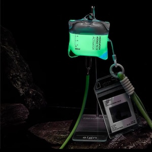 Puede incluir: Una bolsa intravenosa verde brillante con el texto "MEDICATION ADDED TO IV ACTIVATOR" y una medida de 50 ml. Un tarjetero de plástico transparente con un cordón verde y un soporte metálico con la palabra "sign" también son visibles.