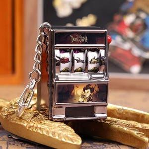 May include: A silver-colored slot machine keychain with the words "Don't Starve" on the top. The slot machine has three reels with various images and a small red lever. The bottom of the machine features a cartoon image.
