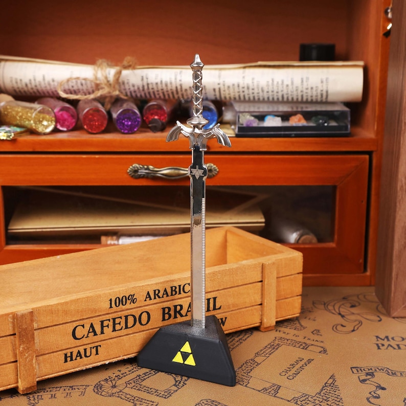 May include: A silver-colored sword figurine with a black base featuring a yellow triangle design. The sword has intricate detailing on the hilt and blade. The figurine is displayed on a wooden surface.
