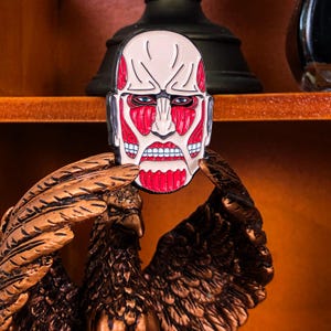 Close-up shot of the Colossal Titan enamel pin against a wooden tabletop background, capturing the intricate muscle texture and sharp facial expression from the anime series Attack on Titan. The photo highlights the clean lines and bold outlines filled with rich red enamel, demonstrating its craftsmanship. Ideal for use as a backpack pin, denim jacket accent, or collectible item for anime and manga lovers. The lighting in the image enhances the pin's gloss and texture.