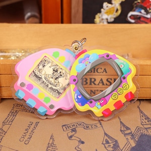 May include: A clear, plastic charm in the shape of a vintage handheld game. The pink side features a gold-colored emblem with the text "HOGWARTS". The yellow side has a screen with the text "SICA BRAS" and colorful buttons. A silver-colored ring is at the top.