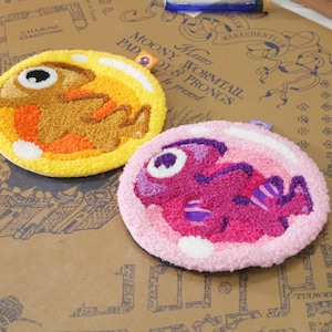 Pode incluir: Duas bases redondas e tufadas. Uma é amarela com um desenho de criatura marrom, a outra é rosa com um desenho de criatura roxa. Ambas têm detalhes em branco e preto. As bases são feitas à mão e têm um estilo fantasioso e caricatural.