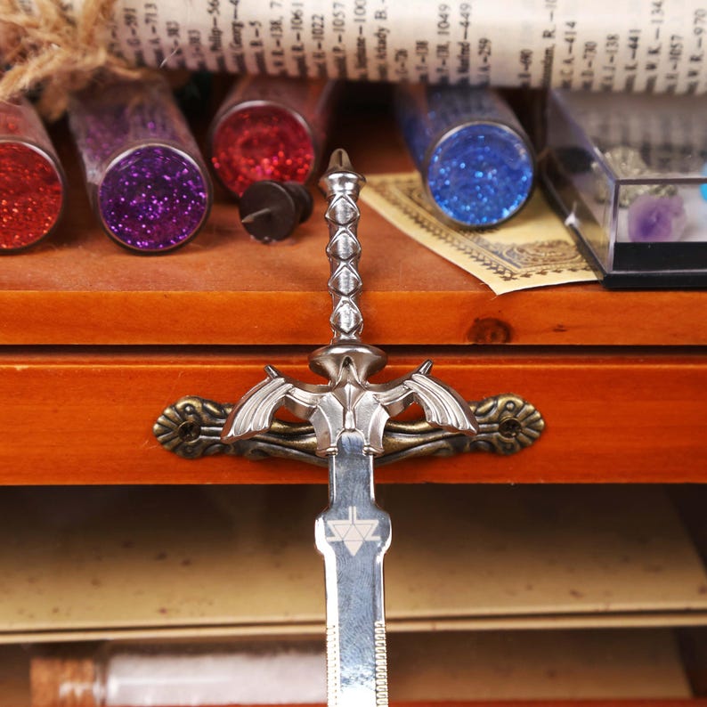 May include: A silver-colored decorative sword with a detailed hilt and blade. The sword is positioned in front of a wooden drawer, with various glass vials filled with colorful glitter and a small box containing gemstones in the background. The sword's design includes a symbol on the blade.