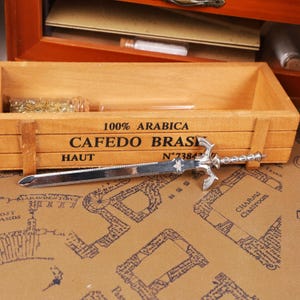 May include: A silver letter opener shaped like a sword, with a decorative hilt and a pointed blade. The opener rests on a map-patterned surface, near a wooden box with the text "100% ARABICA CAFEDO BRAS HAUT N&deg;7384".