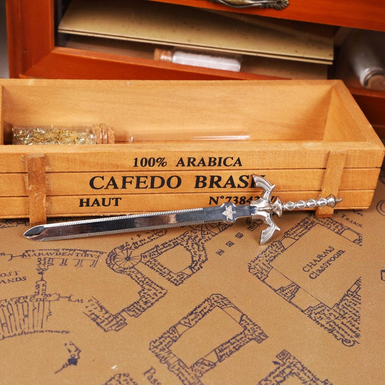 May include: A silver letter opener shaped like a sword, with a decorative hilt and a pointed blade. The opener rests on a map-patterned surface, near a wooden box with the text "100% ARABICA CAFEDO BRAS HAUT N&deg;7384".