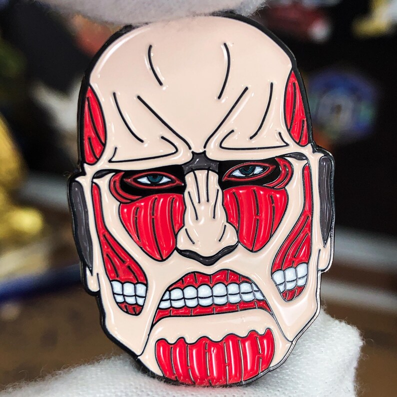 Product display image of the Colossal Titan enamel pin staged beside gaming accessories and collectible anime merchandise, showing its relevance for platform gamers, collectors, and fans of Attack on Titan. The pin stands out with bold red coloring and stylized features of the titan’s face. Designed for jackets, bags, and holiday gifts, the pin is shown in an environment that appeals to its target audience of anime and gaming culture enthusiasts.