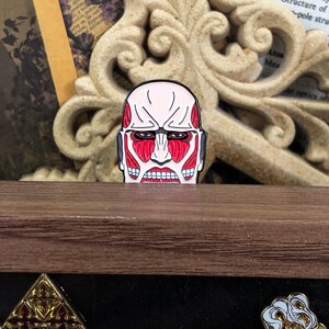 Colossal Titan enamel pin placed on a denim jacket lapel to demonstrate its wearable size and pop culture fashion appeal. The red and black titan design contrasts with the blue fabric, showcasing its striking appearance. The pin’s secure rubber clutch is not visible from this angle, giving it a seamless accessory look. This product image is targeted at anime fans who enjoy customizing streetwear and accessories with themed merchandise and collectible items.