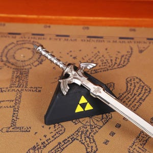 Full set display of the Zelda Link Master Sword Letter Opener with Triforce base, placed stylishly on a wooden surface for a modern collectible look. This functional miniature sword is ideal for opening letters or packages, while serving as fantasy desk decor and a gamer conversation piece. A must-have holiday gift idea for fans of Tears of the Kingdom, Breath of the Wild, and Ocarina of Time, combining nostalgia with practicality for birthdays, Christmas, and Halloween.
