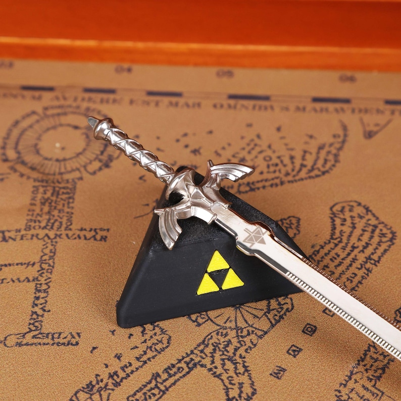 Full set display of the Zelda Link Master Sword Letter Opener with Triforce base, placed stylishly on a wooden surface for a modern collectible look. This functional miniature sword is ideal for opening letters or packages, while serving as fantasy desk decor and a gamer conversation piece. A must-have holiday gift idea for fans of Tears of the Kingdom, Breath of the Wild, and Ocarina of Time, combining nostalgia with practicality for birthdays, Christmas, and Halloween.