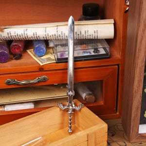 May include: A silver-colored miniature sword with a detailed hilt and blade. The sword is standing upright in front of a wooden box and a wooden cabinet with drawers. The background includes small glass jars and a rolled-up paper.