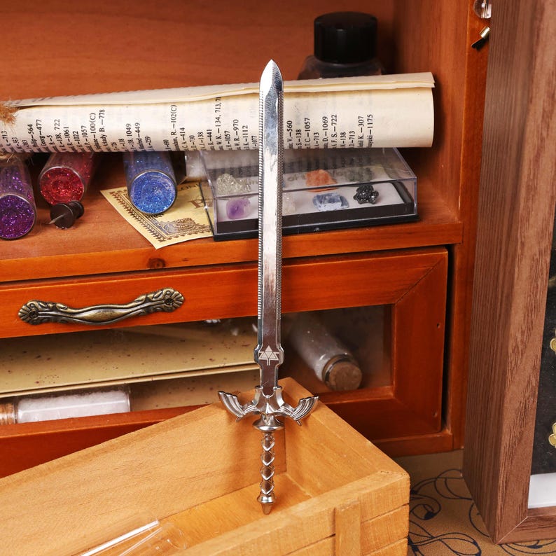 May include: A silver-colored miniature sword with a detailed hilt and blade. The sword is standing upright in front of a wooden box and a wooden cabinet with drawers. The background includes small glass jars and a rolled-up paper.