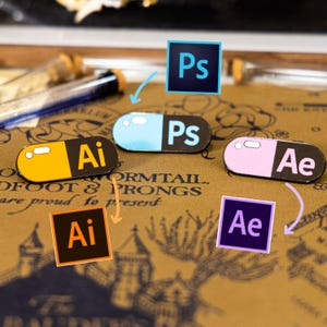 May include: Enamel pins featuring Adobe software logos: Ai (Illustrator), Ps (Photoshop), and Ae (After Effects). The pins are designed as capsules in yellow, blue, and pink, with the software initials displayed. The background is a textured, sepia-toned surface.