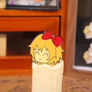 May include: A decorative candle with a cartoon character topper. The topper features a smiling face with yellow hair and a red bow. The candle is white with a dripped wax design.