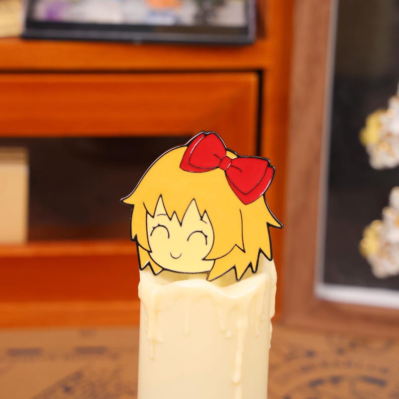May include: A decorative candle with a cartoon character topper. The topper features a smiling face with yellow hair and a red bow. The candle is white with a dripped wax design.
