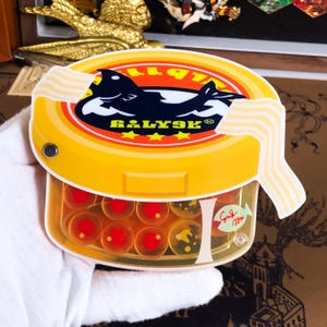 Op de afbeelding: Een gele en oranje plastic container met een zwart-witte visillustratie op de deksel. De deksel heeft de tekst "Balyak" en een witte streep rond de rand. De container is gevuld met kleine, ronde, oranje en rode snoepjes.