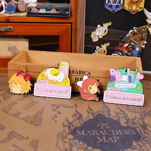 May include: A collection of colorful anime-style enamel pins. The pins feature characters and phrases like "I want breakfast" and "Welcome to the club!" The pins are set against a wooden box and a map background.