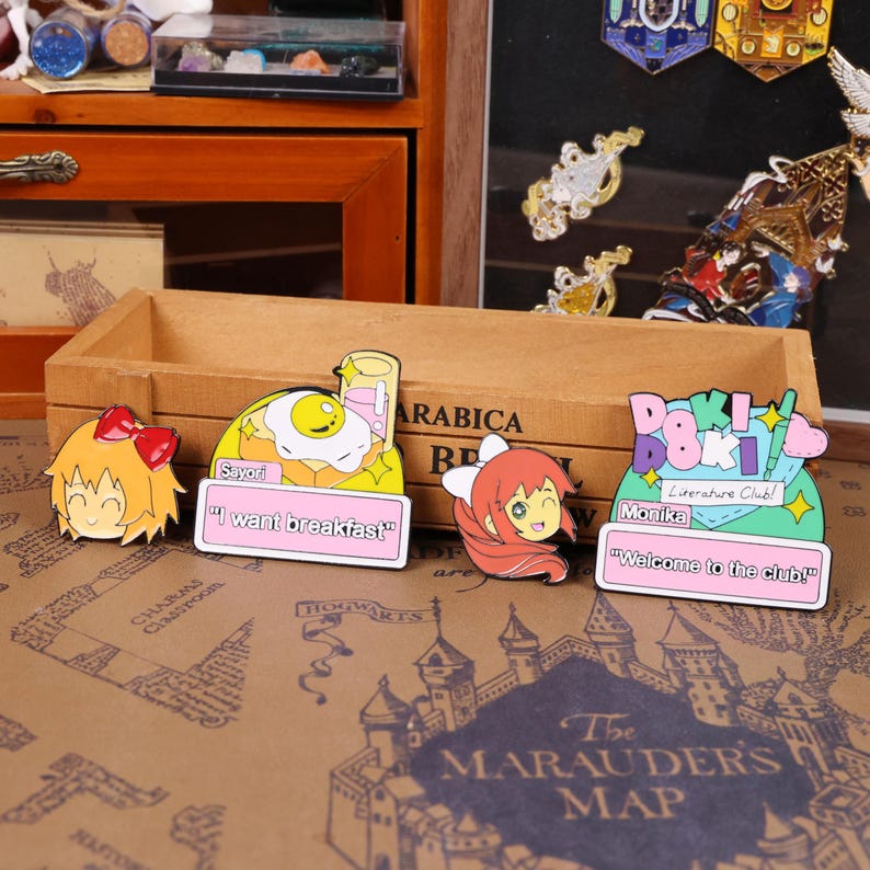May include: A collection of colorful anime-style enamel pins. The pins feature characters and phrases like "I want breakfast" and "Welcome to the club!" The pins are set against a wooden box and a map background.
