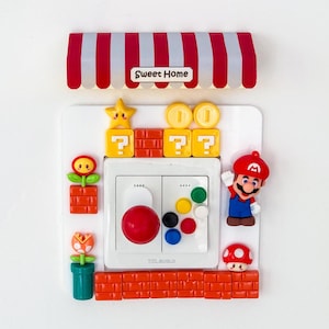 Puede incluir: Una cubierta de interruptor de luz blanca decorada con elementos temáticos de Super Mario Bros. Incluye un toldo a rayas rojas y blancas con el texto "Sweet Home", una figura de Mario, bloques de interrogación, monedas y un hongo.