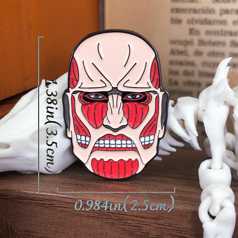 Attack on Titan Colossal Titan enamel pin lying flat on a white fabric surface to emphasize its bold design and compact size. The pin depicts the titan's signature muscular face in bright red with detailed linework and expressive anime-style features. The rubber clutch backing is visible, hinting at the secure fit for everyday use. The pin’s polished surface reflects light slightly, drawing attention to the quality of its materials and vibrant enamel fill.