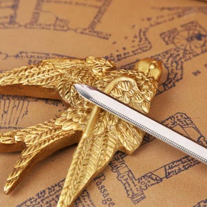 Zelda Master Sword Letter Opener displayed on a wooden desk with its Triforce base stand, showcasing the polished metal blade and detailed hilt. This image highlights the fantasy-inspired design of the letter opener, perfect as a Zelda fan collectible, Christmas gamer gift, or geek office decor piece. Ideal for fans of Tears of the Kingdom, BOTW, and Ocarina of Time, this functional mini sword adds a magical touch to any holiday gift set, Halloween display, or stocking stuffer collection.