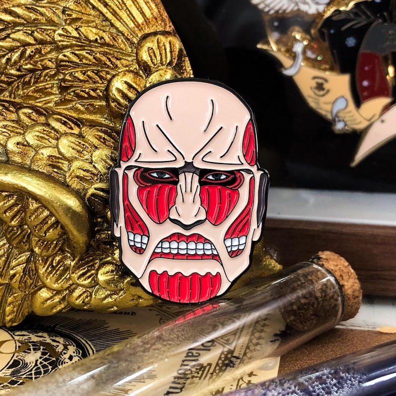 Colossal Titan enamel pin featuring intense red muscle detailing and bold black outline, inspired by the iconic character from Attack on Titan anime. The pin is shown up close to highlight the vivid color contrast and fine craftsmanship of the zinc alloy with soft enamel finish. Ideal for anime fans, cosplayers, and collectors, the pin is designed to be worn on jackets, bags, or displayed with other fandom collectibles. Photographed on a neutral background to showcase design clarity.