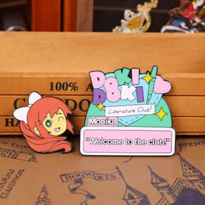 May include: Two enamel pins featuring anime-style characters. One pin shows a winking girl with red hair and a white bow. The other pin has the text "Doki Doki Literature Club!" and "Welcome to the club!" on a pink background.
