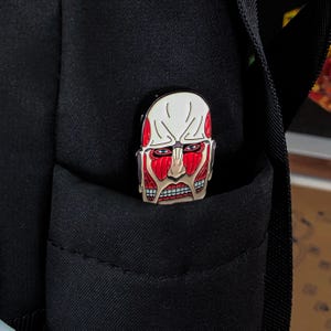 Product display image of the Colossal Titan enamel pin staged beside gaming accessories and collectible anime merchandise, showing its relevance for platform gamers, collectors, and fans of Attack on Titan. The pin stands out with bold red coloring and stylized features of the titan’s face. Designed for jackets, bags, and holiday gifts, the pin is shown in an environment that appeals to its target audience of anime and gaming culture enthusiasts.