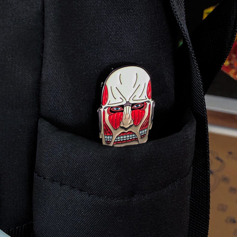 Product display image of the Colossal Titan enamel pin staged beside gaming accessories and collectible anime merchandise, showing its relevance for platform gamers, collectors, and fans of Attack on Titan. The pin stands out with bold red coloring and stylized features of the titan’s face. Designed for jackets, bags, and holiday gifts, the pin is shown in an environment that appeals to its target audience of anime and gaming culture enthusiasts.