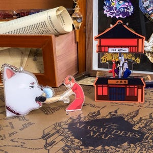Könnte beinhalten: Zwei Acryl-Anhänger mit Figuren aus dem Anime Gintama. Der erste Anhänger ist ein weißer Hund mit roten Augen und einer herausgestreckten rosa Zunge. Der zweite Anhänger ist ein rotes Gebäude mit einem Schild, auf dem "万事屋銀ちゃん" in japanischer Sprache steht.