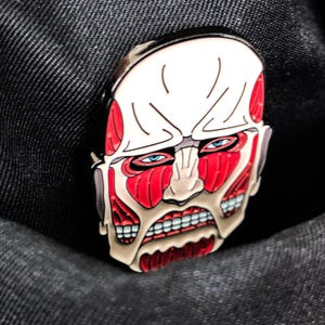 Close-up shot of the Colossal Titan enamel pin against a wooden tabletop background, capturing the intricate muscle texture and sharp facial expression from the anime series Attack on Titan. The photo highlights the clean lines and bold outlines filled with rich red enamel, demonstrating its craftsmanship. Ideal for use as a backpack pin, denim jacket accent, or collectible item for anime and manga lovers. The lighting in the image enhances the pin's gloss and texture.