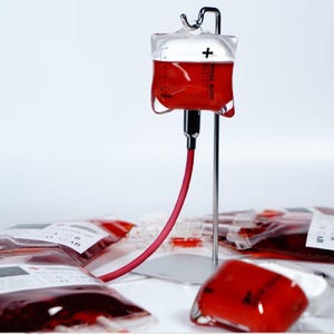 Può includere: Una sacca di sangue in vetro riempita con un liquido rosso, sospesa a un supporto metallico. La sacca ha una parte superiore bianca con un segno più e un tubo rosso. Diverse sacche simili sono visibili sullo sfondo.