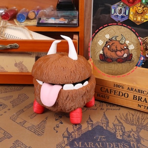 May include: A whimsical brown monster figurine with white horns, a pink tongue, and red feet. The figurine has a furry texture and an open mouth. A circular illustration of a similar monster is also visible.