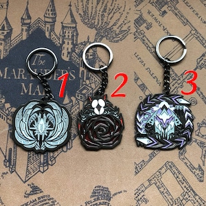 Può includere: Tre portachiavi in metallo nero con diversi design. Il primo portachiavi presenta un design bianco e argento con una creatura alata. Il secondo portachiavi presenta un design di rosa rossa e nera. Il terzo portachiavi presenta un design viola e argento con una creatura alata.