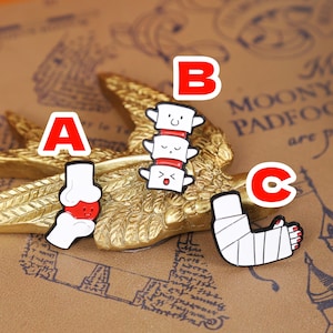 May include: Three medical-themed enamel pins. Pin A shows a knee joint, pin B depicts a spine, and pin C features a bandaged arm with red nail polish. Each pin has a white outline and a cartoonish design.