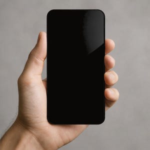 May include: A black smartphone with rounded corners is held in a person's hand. The phone's screen is completely black, reflecting light. The background is a plain, light gray.