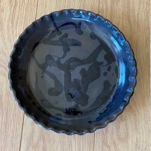 Puede incluir: Un plato para tartas de cerámica negra con un borde festoneado y un glaseado azul oscuro. El glaseado tiene un efecto moteado con remolinos negros y azul oscuro.