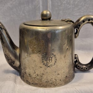 Könnte beinhalten: Eine silberne Teekanne mit einer angelaufenen Oberfläche und einem Monogramm. Die Teekanne hat einen runden Deckel und einen gebogenen Griff.