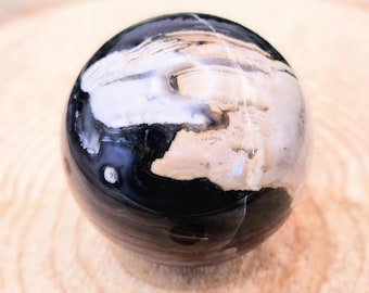 May include: A polished sphere with a mix of black, white, and tan colors. The orb has a glossy finish and appears to be made of stone or a similar material. The sphere is sitting on a light brown wooden surface.