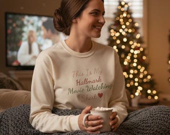 May include: Cream-colored sweatshirt with the text "This Is My Hallmark Movie Watching Shirt" in pink, green, and red. A heart symbol is below the text. The sweatshirt is worn by a person holding a mug.
