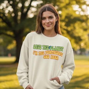 Może przedstawiać: Kremowa bluza z napisem "SAVE THE PLANET I'M THE FUNNIEST ONE HERE" w kolorze zielonym, niebieskim, żółtym i pomarańczowym. Bluza jest noszona przez osobę w plenerze z rozmytym zielono-żółtym tłem.