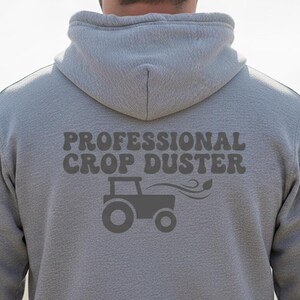 May include: A gray hooded sweatshirt with the text "PROFESSIONAL CROP DUSTER" and a tractor graphic. The design is in a darker gray color. The sweatshirt is made of a soft material.
